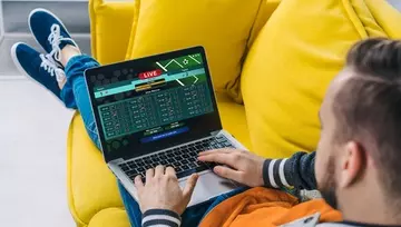 a man relaxing on a yellow sofa placing a sports bet on his laptop