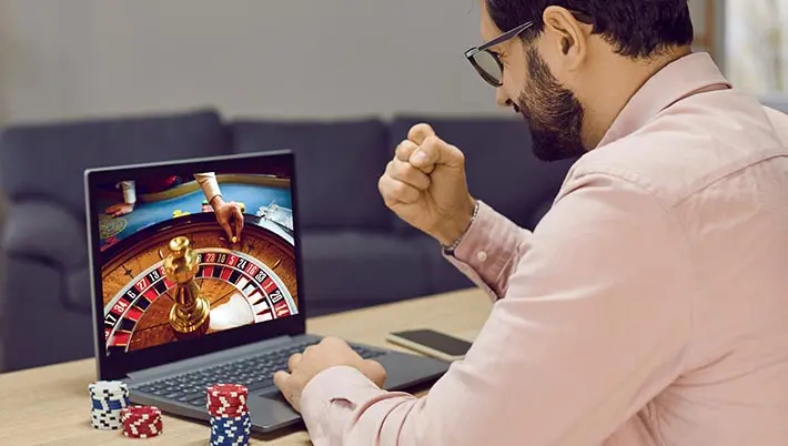 man looking at a screen with a roulette wheel, making a “winning” gesture