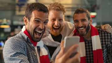 Three men wearing team scarves excitedly watching their team win on their mobile phone