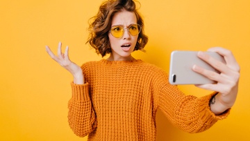 young woman in yellow sweater confused looking at her mobile phone on a yellow background
